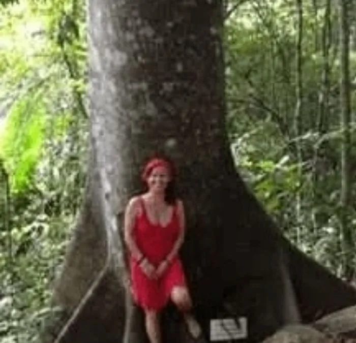 Isa Herrera in a red dress seated against the buttress roots of a rainforest tree.