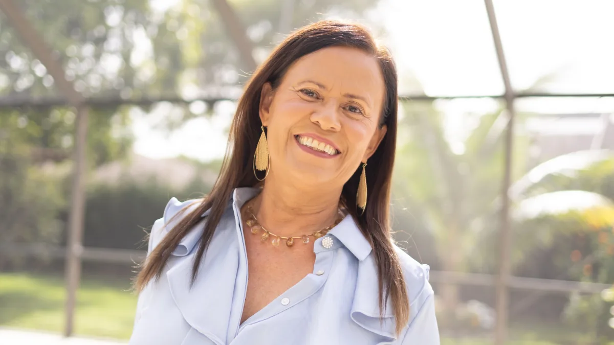 Isa Herrera smiling in a pale blue shirt outdoors, backlit by sun through a screened garden.