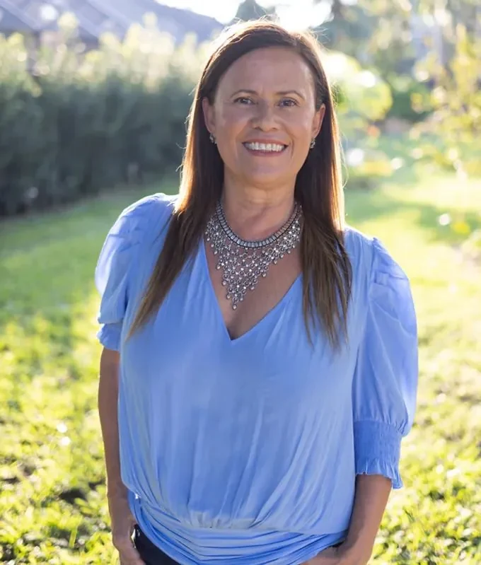 Isa Herrera in a blue blouse and silver statement necklace, standing outdoors on sunlit grass.